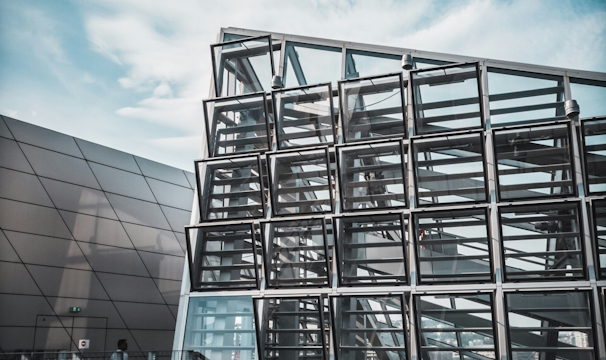A modern architectural structure composed of a geometric grid of glass panels reflecting the ambient sky. The design features overlapping metal and glass elements situated in an urban environment. The image captures a sense of modernity and innovation.